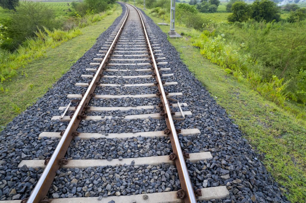 Die Bahngleise schlängeln sich durch eine grüne Landschaft, gesäumt von Büschen und Bäumen, wobei der Boden zwischen und um die Schienen herum mit Kies bedeckt ist.