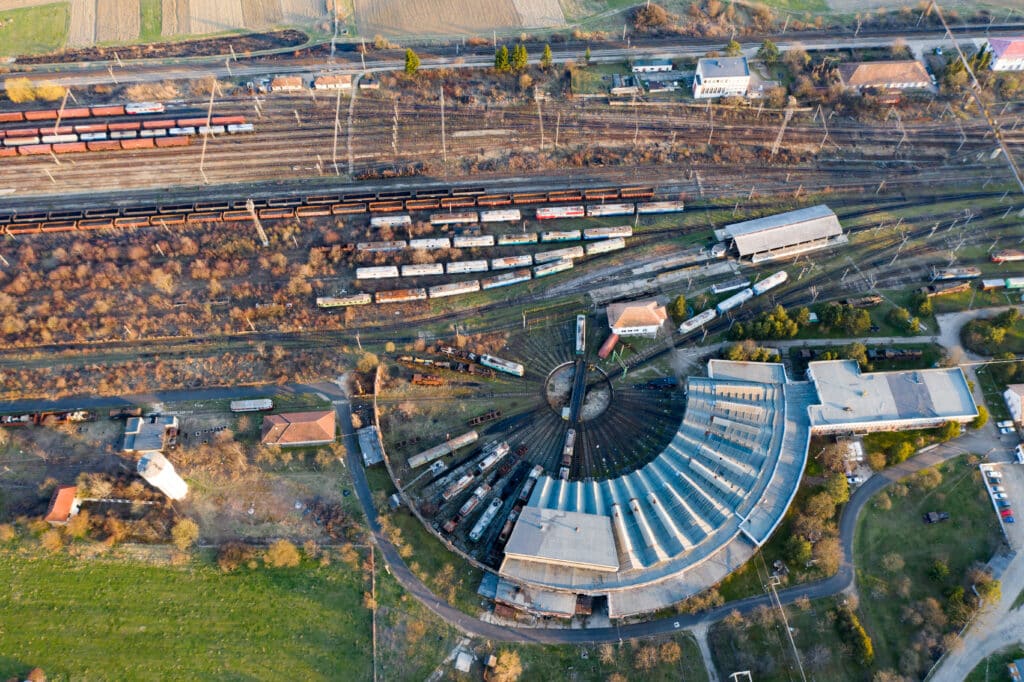 Aerial view of a train roundhouse with multiple railway tracks, trains, and surrounding buildings, set in a partially grassy and partially wooded area.