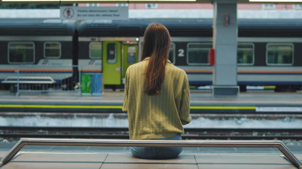 Eine Frau mit langen braunen Haaren sitzt auf einer Bank an einem Bahnhof, den Rücken zur Kamera gewandt, und blickt auf einen stehenden Zug auf dem gegenüberliegenden Bahnsteig.