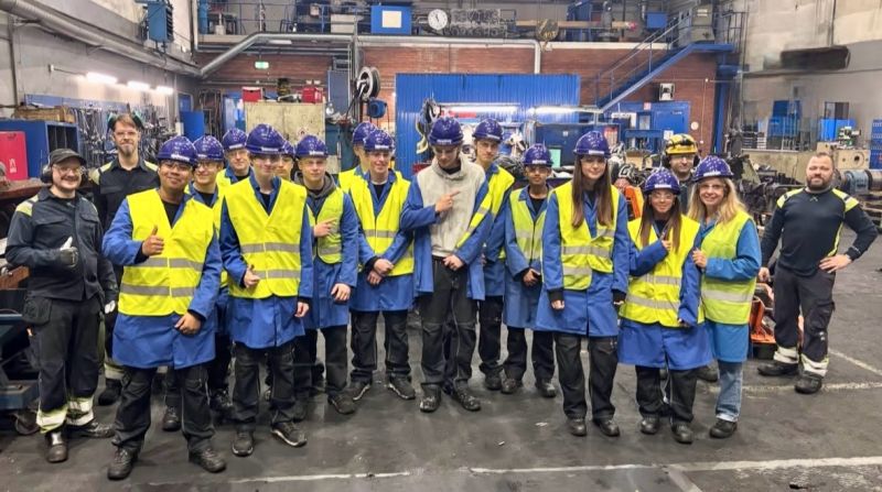 Eine große Gruppe von Schülern, die blaue Schutzhelme und gelbe Warnwesten tragen, posiert in einer Maschinenwerkstatt für ein Foto.