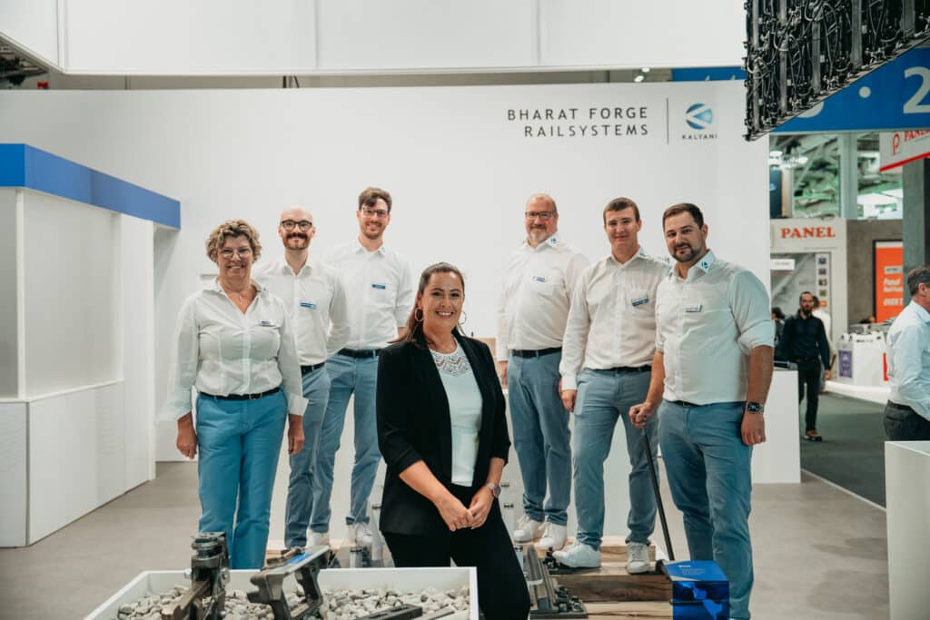 Eine Gruppe von sieben Personen in weißen Hemden und Business-Casual-Kleidung posiert für ein Foto vor dem Messestand von Bharat Forge Railsystems.